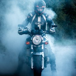 RafalSaduraFotograf.pl - Motocyklista w kasku i kombinezonie na motocyklu, otoczony dymem, ujęcie z przodu, widoczny reflektor i przednie koło.