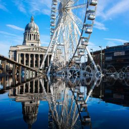 RafalSaduraFotograf.pl - Panorama miejska z budynkiem z kolumnami i diabelskim młynem odbijającymi się w wodzie fontanny w słoneczny dzień.