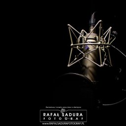 RafalSaduraFotograf.pl - Mikrofon studyjny Neumann w koszyku antywibracyjnym, oświetlony punktowo na czarnym tle, widoczne logo firmy.