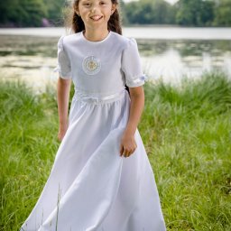 RafalSaduraFotograf.pl - Uśmiechnięta dziewczynka w białej sukience komunijnej z symbolem IHS, stojąca na tle jeziora i zielonej trawy.