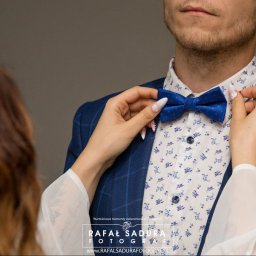 RafalSaduraFotograf.pl - Panna młoda poprawia granatową muchę w czerwone kropki panu młodemu w Gdańsku, który ma na sobie niebieską marynarkę w kratę i koszulę w drobne kwiatki.