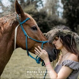RafalSaduraFotograf.pl - Kobieta z wiankiem na głowie dotyka nosem pyska brązowego konia z niebieskim kantarem ozdobionego trawą.