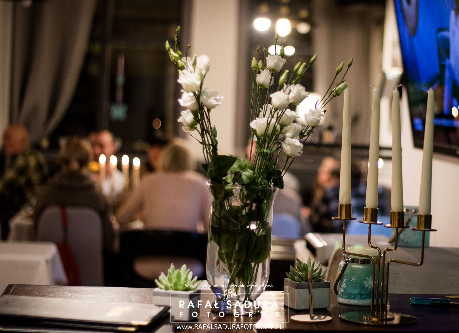 Dekoracja stołu z białymi kwiatami w szklanym wazonie, świecznikiem z czterema świecami, małymi sukulentami w szarych doniczkach i menu, w tle rozmyte postacie gości przy stole.