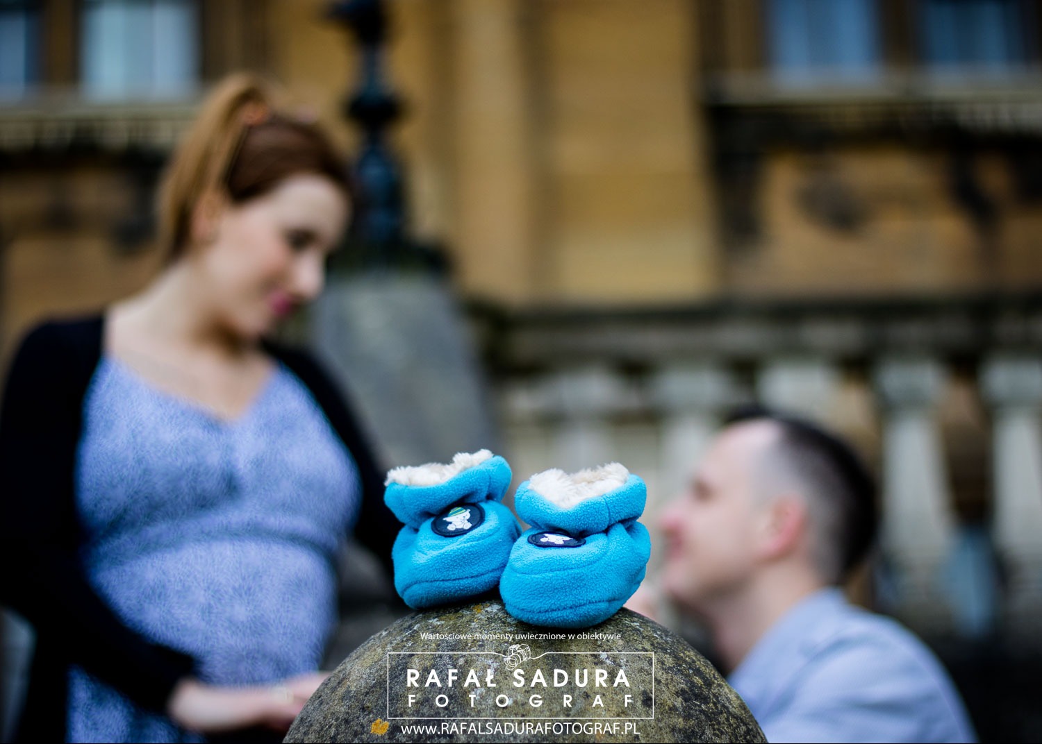 Niebieskie, miękkie buciki dziecięce na kamiennym postumencie, z rozmyte postacie przyszłej mamy i taty w tle, sugerujące sesję ciążową.