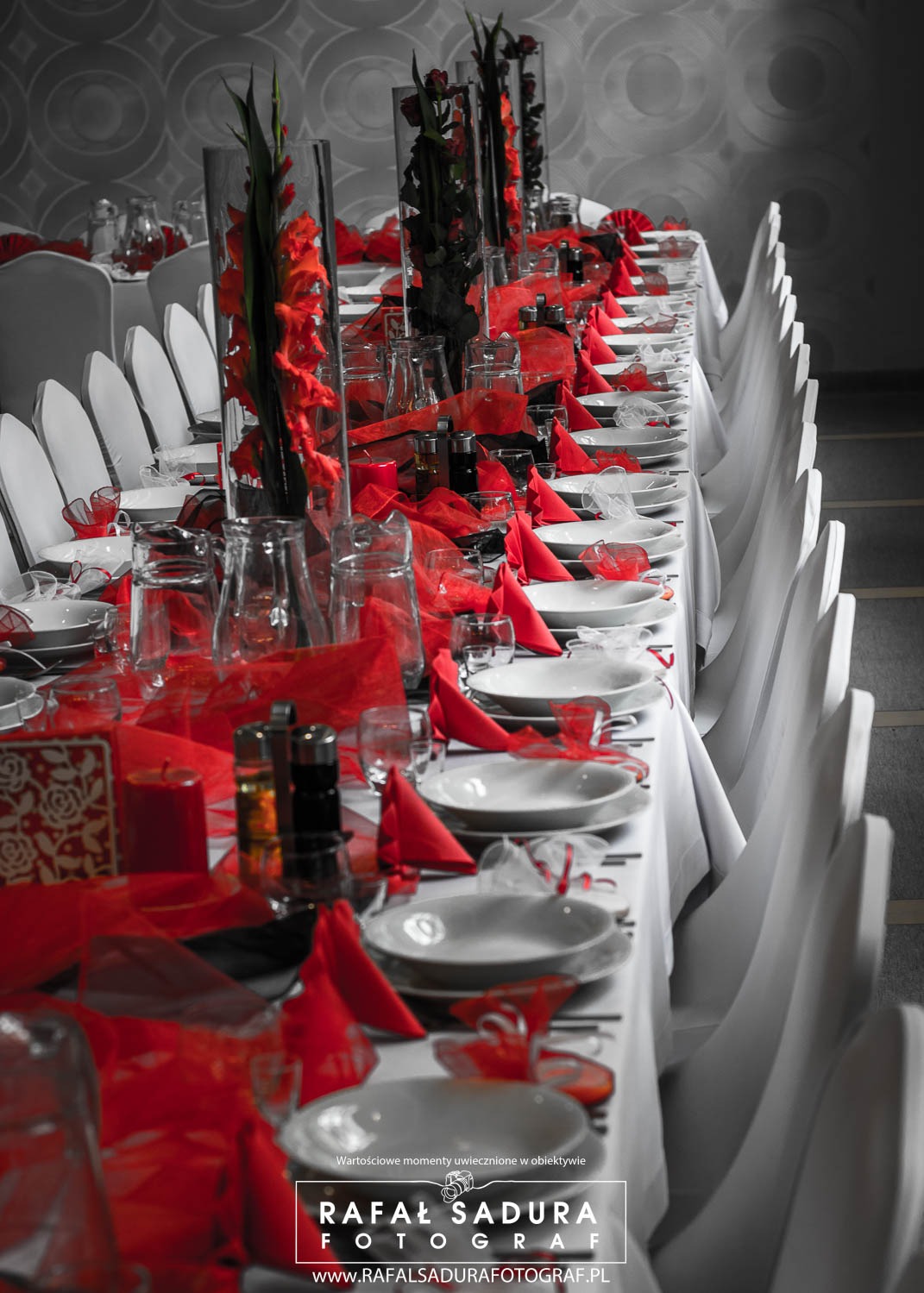 Uroczysty stół weselny z białymi obrusami, czerwonymi serwetkami i kwiatowymi dekoracjami w szklanych wazonach, ustawiony w sali z szarym tłem i białymi krzesłami.