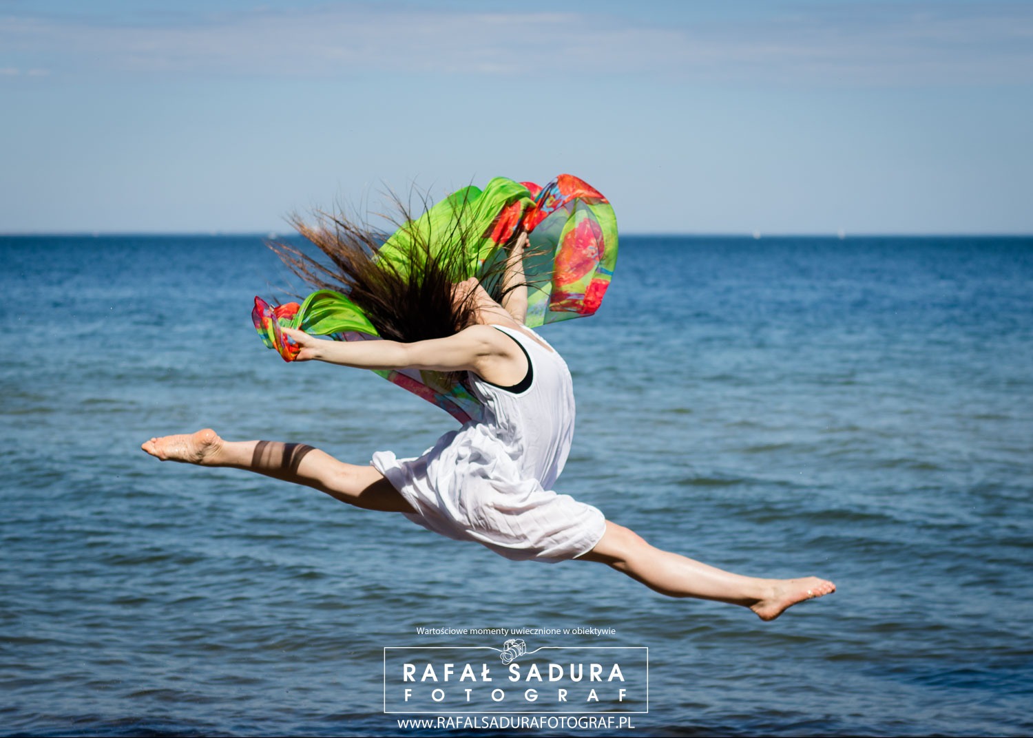Kobieta w białej sukience wykonuje skok w szpagacie na tle morza, z kolorową chustą powiewającą w powietrzu.