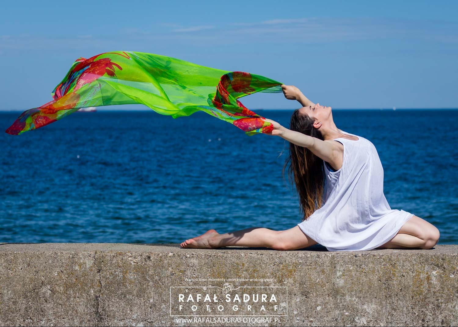 Kobieta w białej sukience siedzi na betonowym murku na tle morza, trzymając w rękach zwiewną, kolorową chustę.