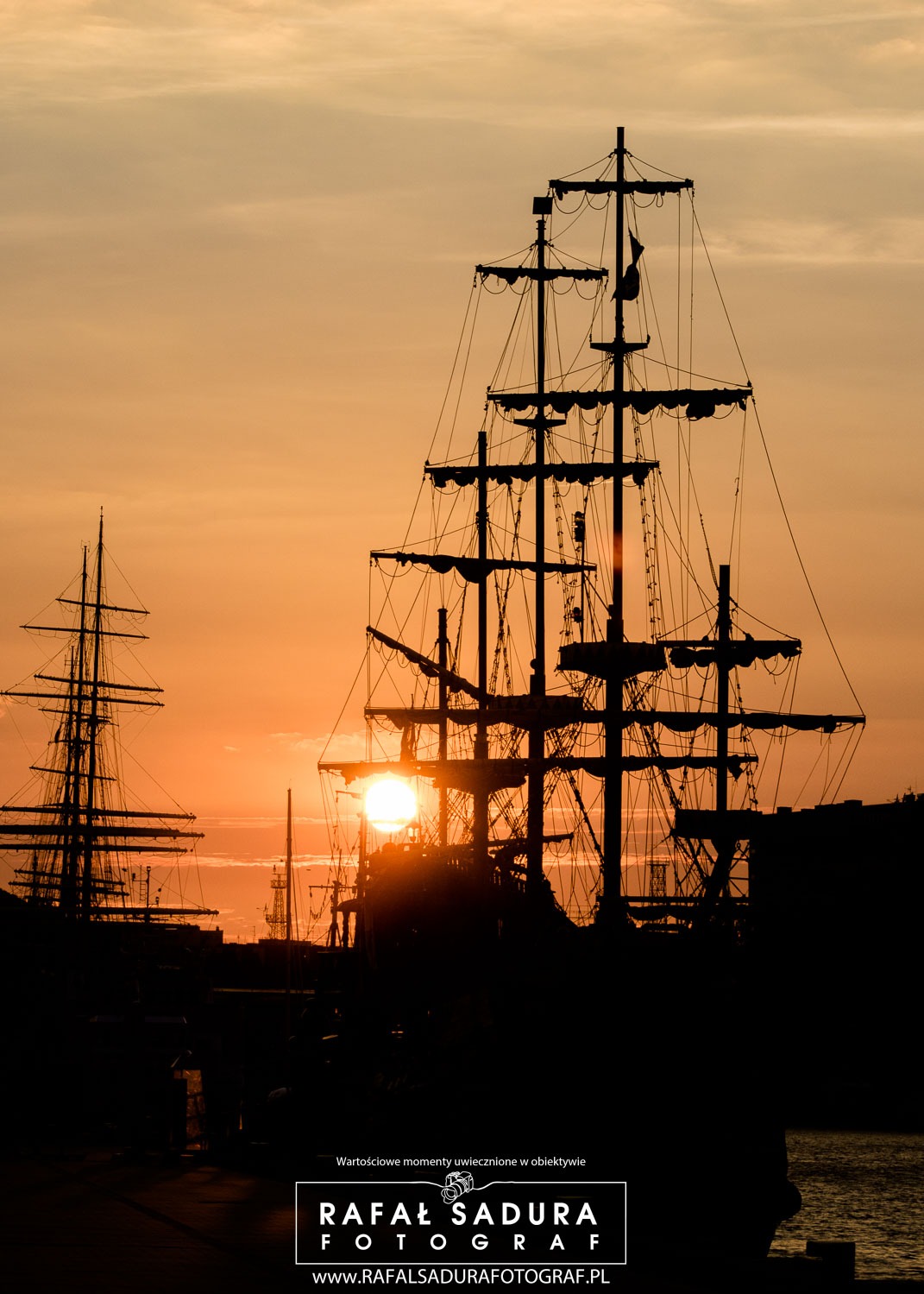 Sylwetki żaglowców na tle pomarańczowego zachodzącego słońca, widoczne maszty, liny i fragmenty kadłubów.