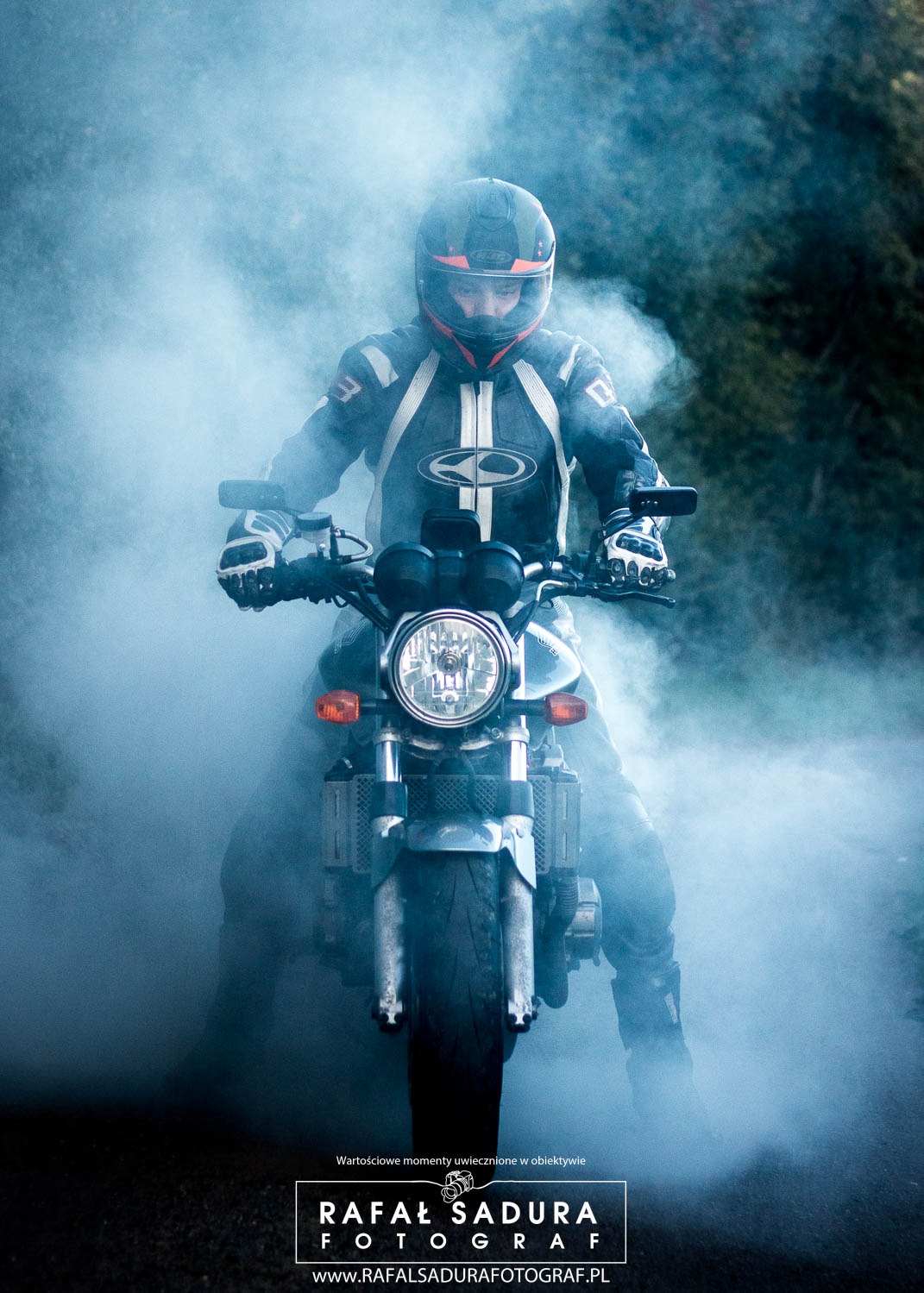 Motocyklista w kasku i kombinezonie na motocyklu, otoczony dymem, ujęcie z przodu, widoczny reflektor i przednie koło.