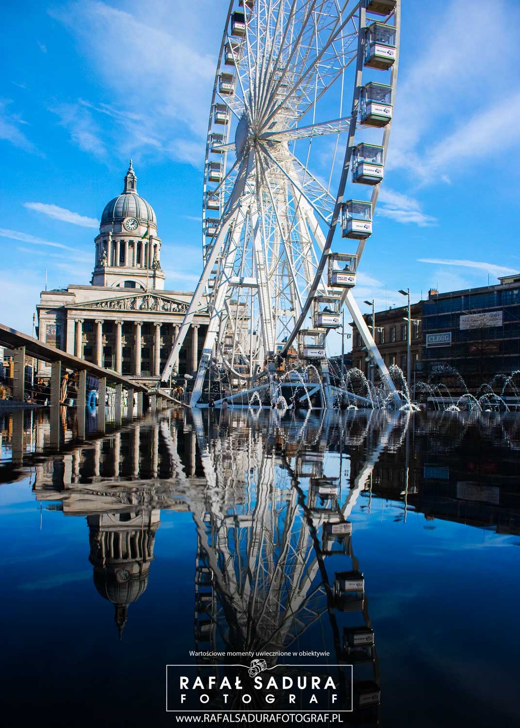 Panorama miejska z budynkiem z kolumnami i diabelskim młynem odbijającymi się w wodzie fontanny w słoneczny dzień.