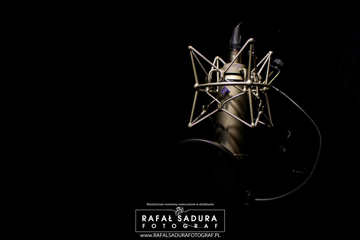 Mikrofon studyjny Neumann w koszyku antywibracyjnym, oświetlony punktowo na czarnym tle, widoczne logo firmy.