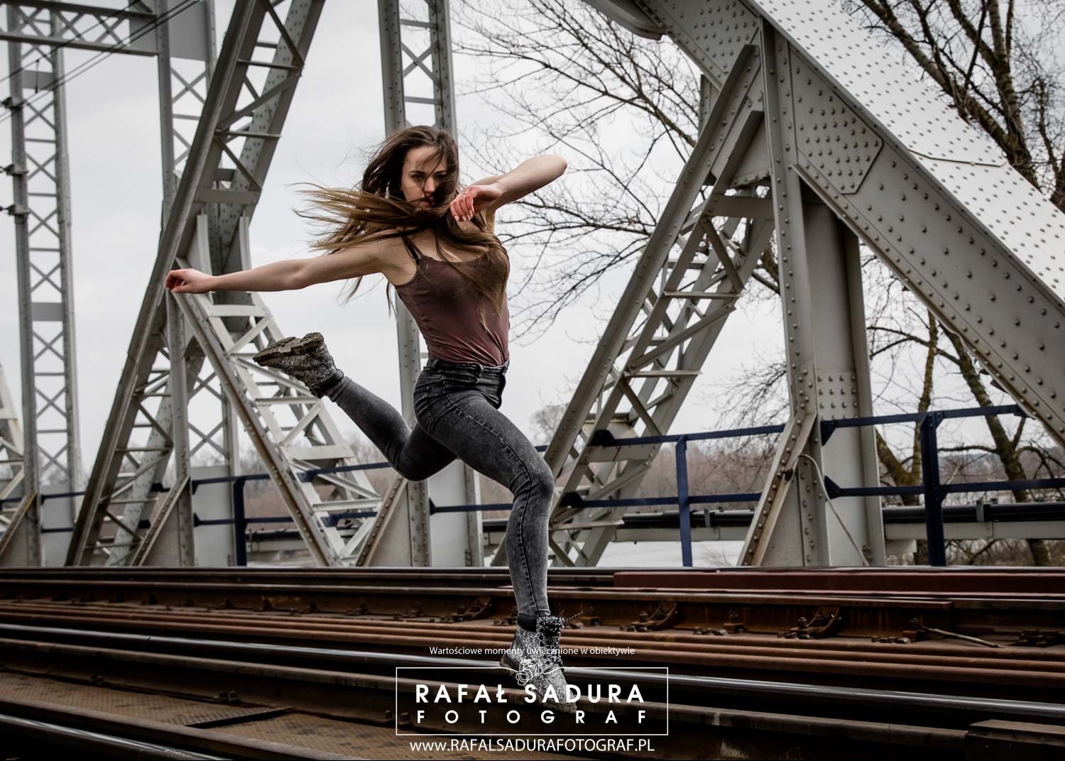 Kobieta w dynamicznym skoku na tle metalowej konstrukcji mostu kolejowego, włosy w ruchu, ujęcie z dołu.