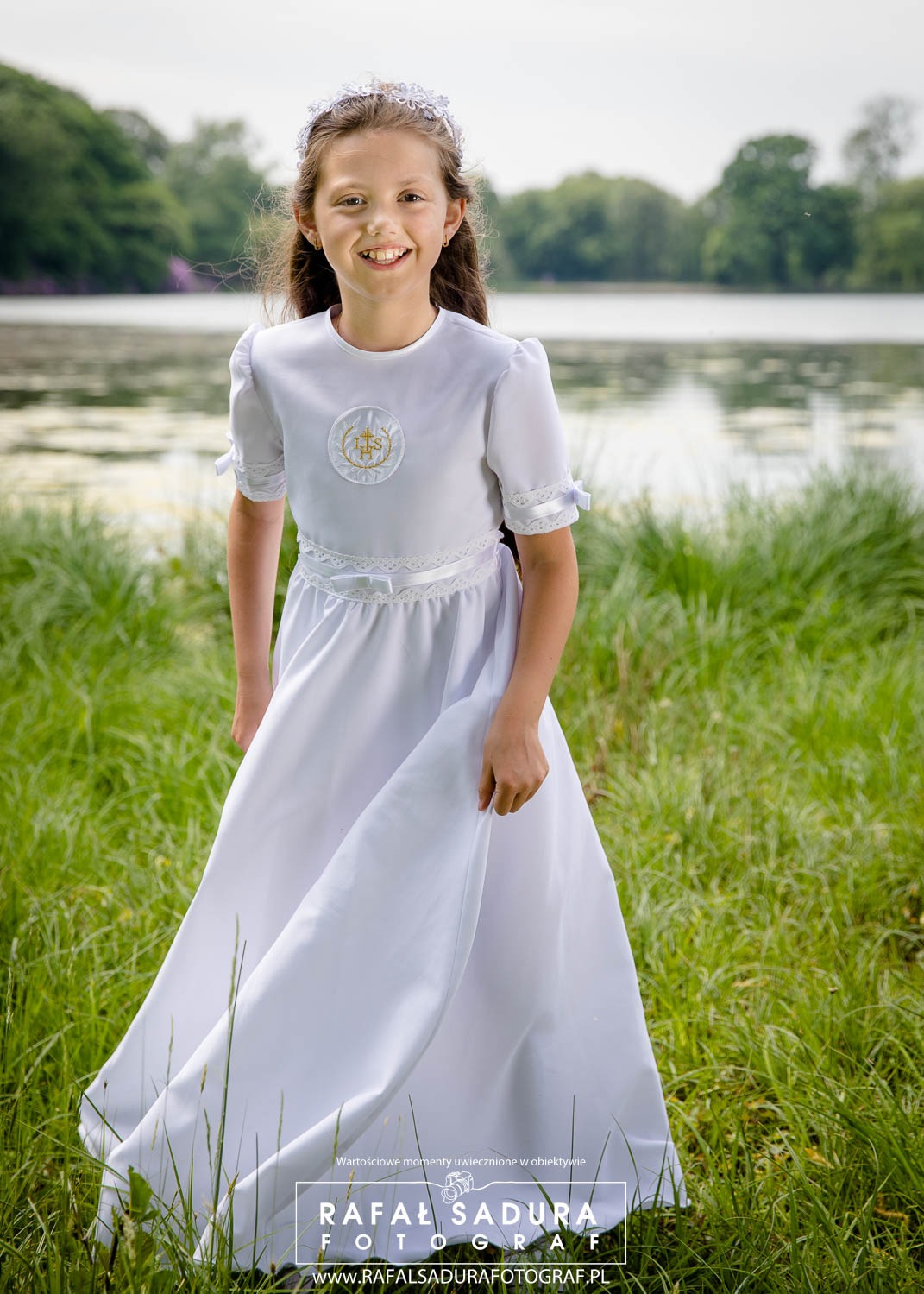 Uśmiechnięta dziewczynka w białej sukience komunijnej z symbolem IHS, stojąca na tle jeziora i zielonej trawy.