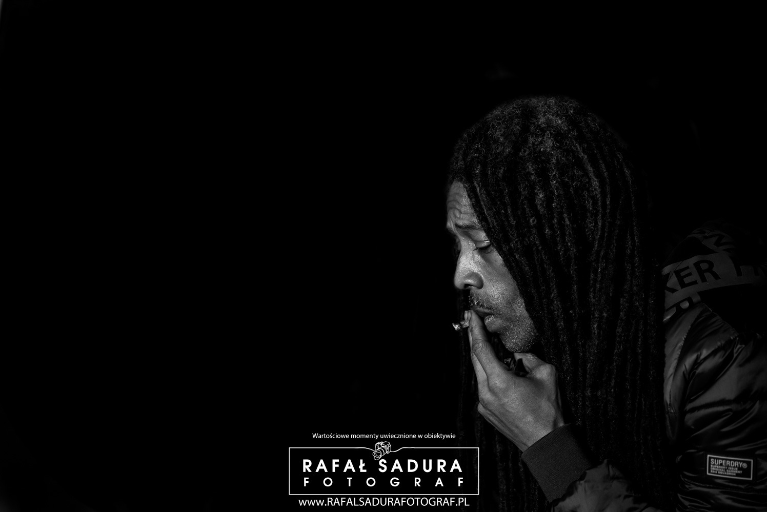 Czarno-biały portret profilowy mężczyzny z dredami, palącego skręta, z ciemnym tłem i widocznym logotypem fotografa w dolnej części kadru.
