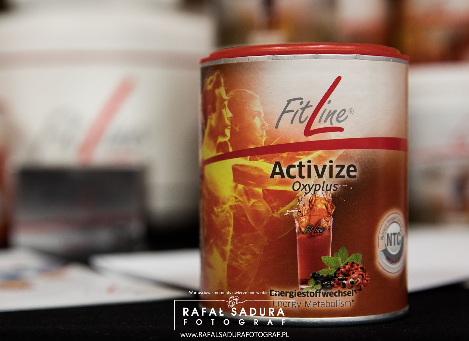 Puszka suplementu diety FitLine Activize Oxyplus z grafiką przedstawiającą sylwetki sportowców i szklankę napoju z owocami, sfotografowana w studiu z rozmytym tłem.