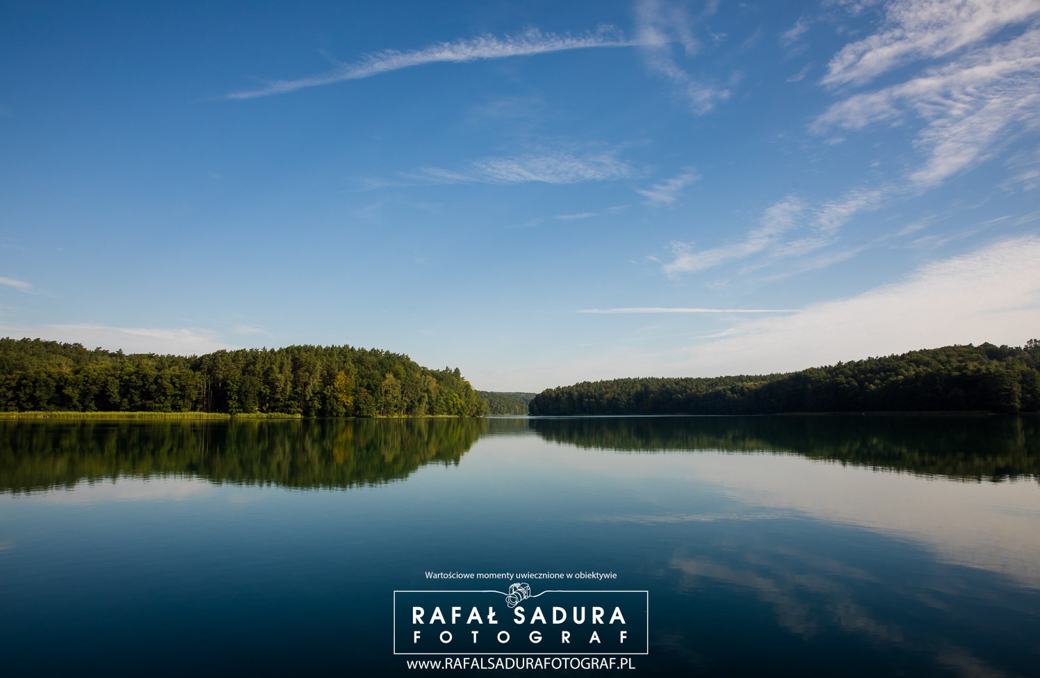 Spokojne jezioro otoczone lasem odbijające się w tafli wody pod błękitnym niebem z delikatnymi obłokami.