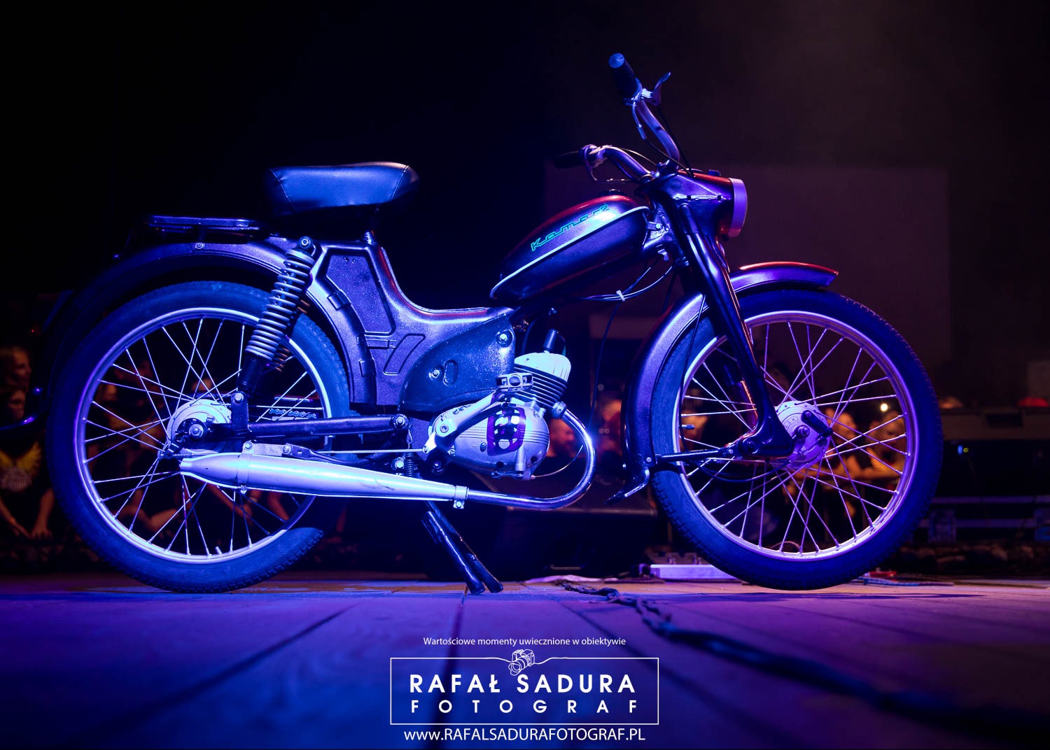 Zdjęcie motoroweru Komar w niebieskim oświetleniu scenicznym, widoczne detale silnika i szprych kół, tło w ciemnych tonacjach z zarysem publiczności.