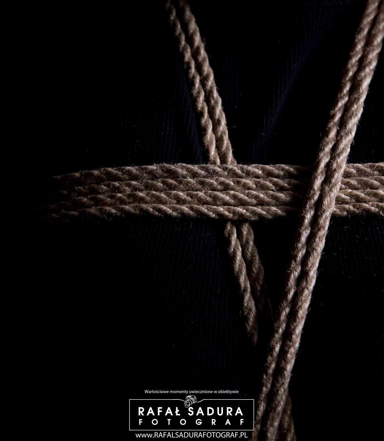 Zbliżenie na dekoracyjne wiązanie z grubego, brązowego sznura na czarnym tle, z widocznym logo fotografa w dolnej części.