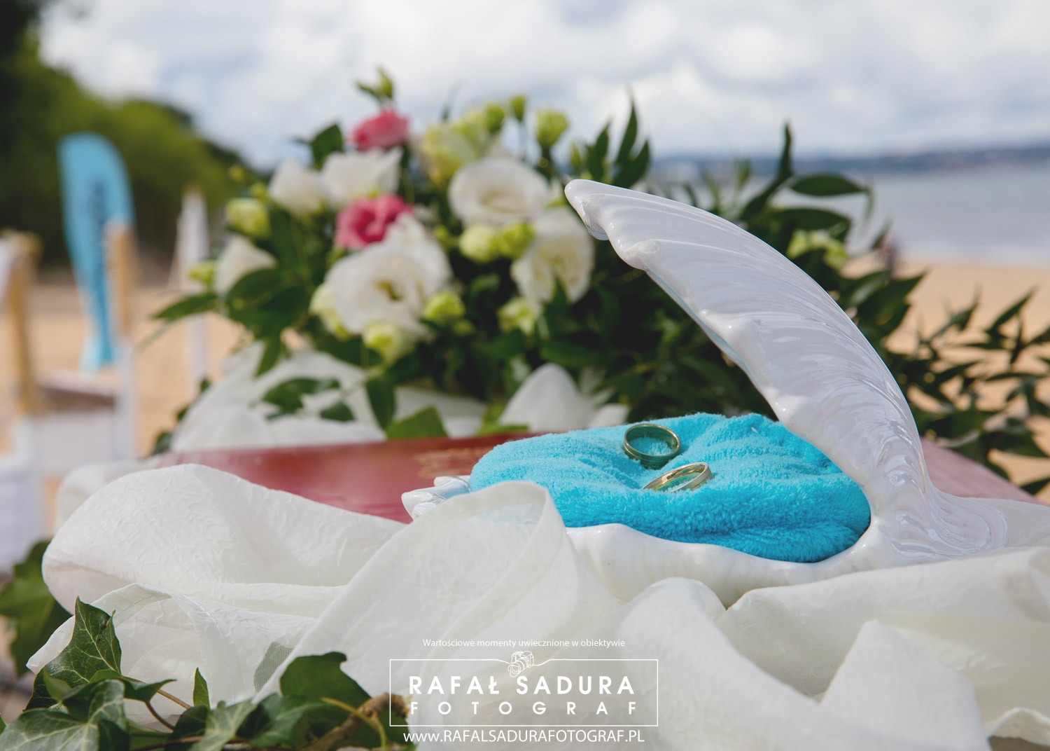 Dwa złote pierścionki ślubne leżące na turkusowej poduszce w białej, ceramicznej muszli, otoczone białymi i różowymi kwiatami na plaży.