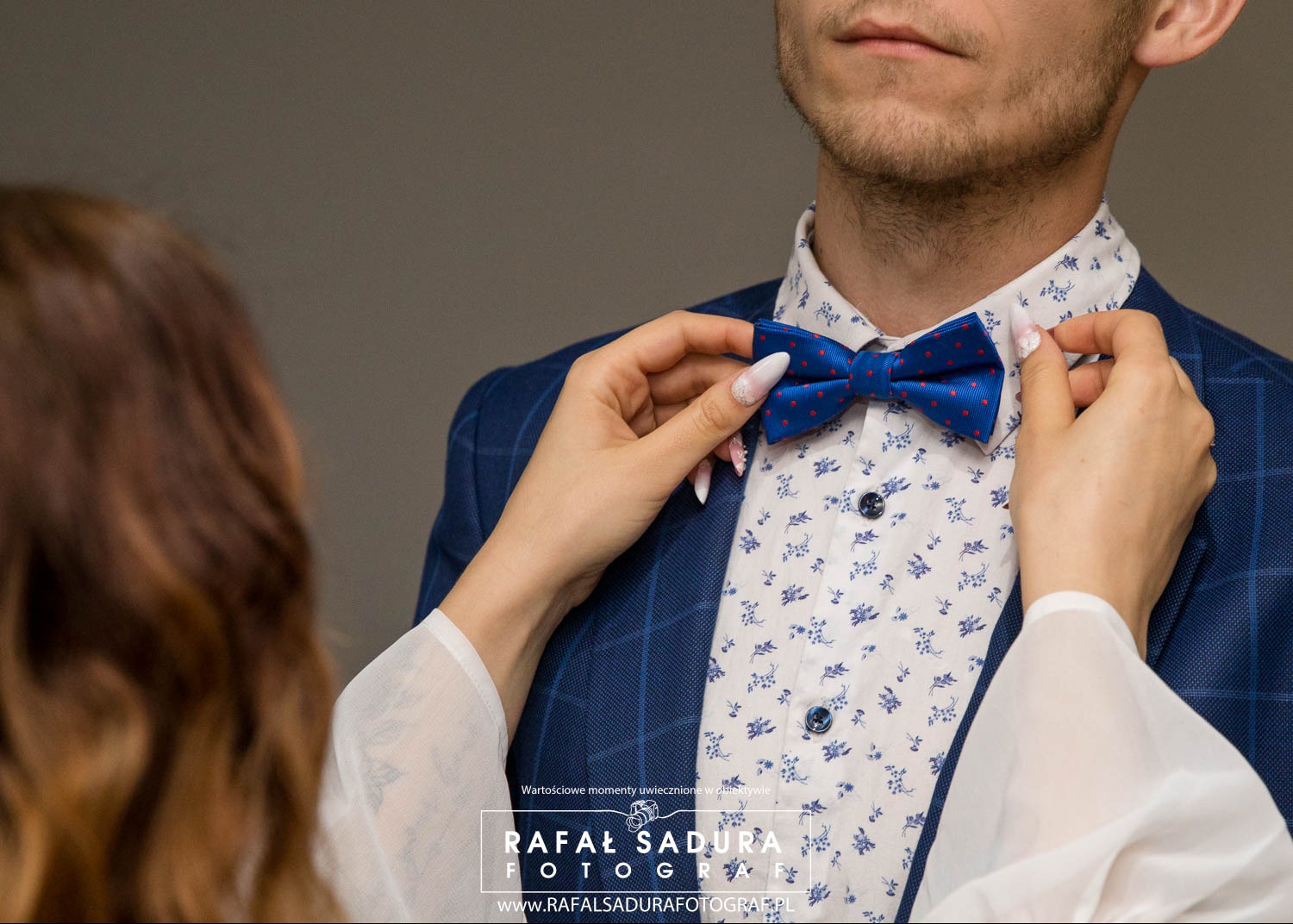 Panna młoda poprawia granatową muchę w czerwone kropki panu młodemu w Gdańsku, który ma na sobie niebieską marynarkę w kratę i koszulę w drobne kwiatki.