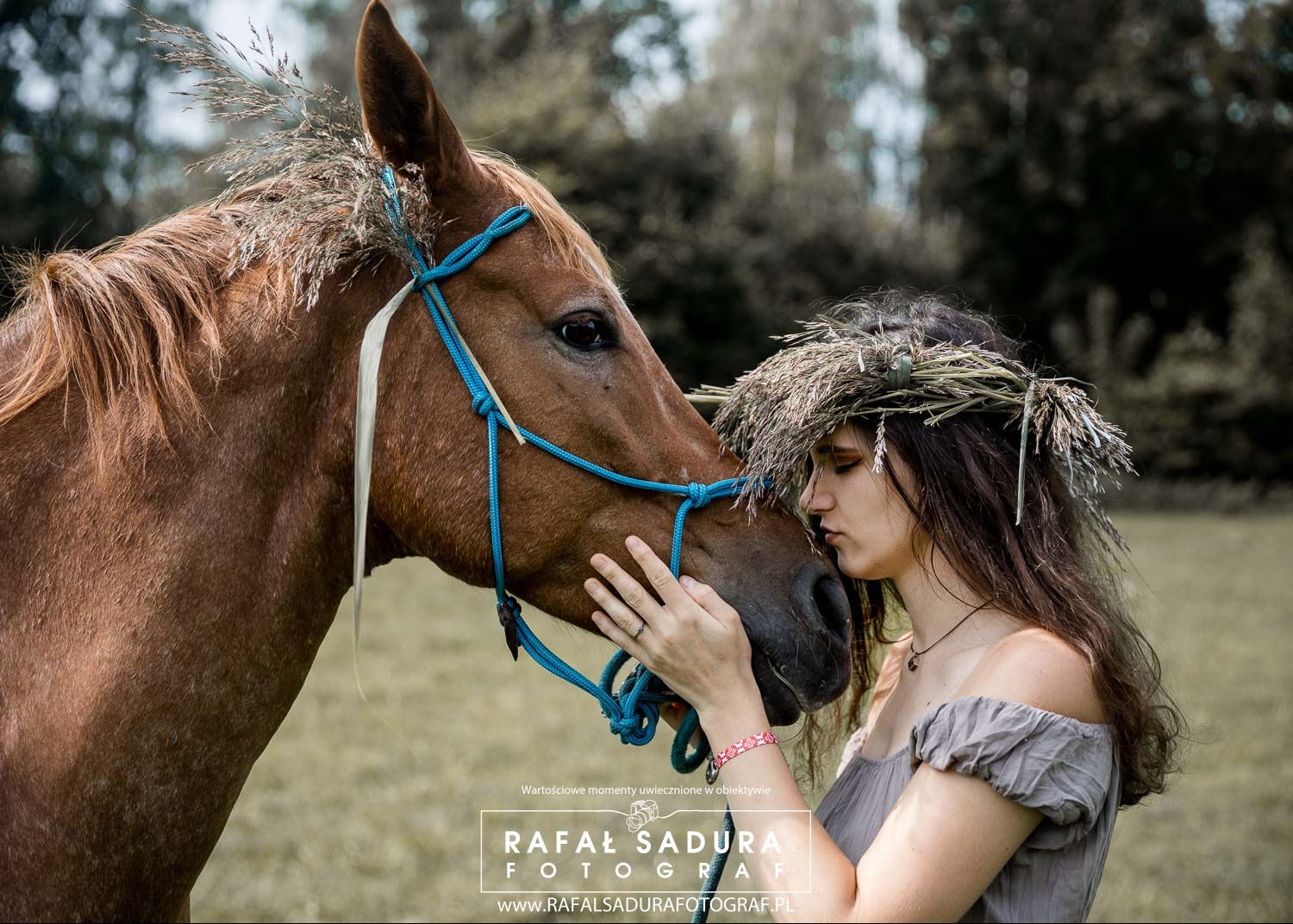 Kobieta z wiankiem na głowie dotyka nosem pyska brązowego konia z niebieskim kantarem ozdobionego trawą.