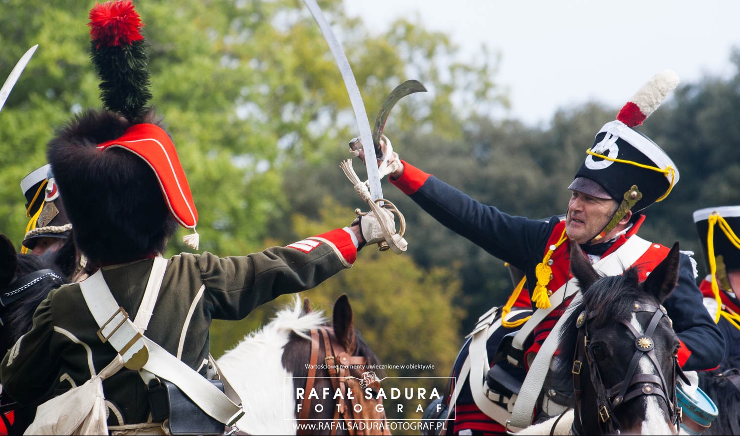 Rekonstrukcja historyczna: Dwóch żołnierzy w mundurach z epoki napoleońskiej na koniach, krzyżujących szable podczas bitwy, z zielonym tłem drzew.