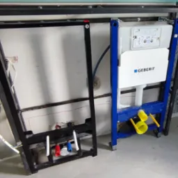 Stelaż podtynkowy Geberit Duofix z widocznymi przyłączami wody (czerwony, niebieski) i odpływu, zamontowany na surowym betonie w trakcie instalacji hydraulicznej.