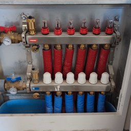 Termogepard sp. z o.o. - Rozdzielacz ogrzewania podłogowego w metalowej szafce, z czerwonymi i niebieskimi rurami, zaworami Ferro i manometrami, widoczne elementy instalacji w szarej izolacji.