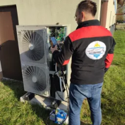 Instalator w okularach, w bluzie z logo firmy, mierzy parametry otwartej obudowy jednostki zewnętrznej klimatyzacji przy użyciu multimetru cyfrowego; obok stoi pompa próżniowa.