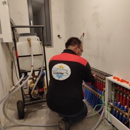 Termogepard sp. z o.o. - Instalator w Miasteczku Śląskim klęczy przed rozdzielaczem ogrzewania podłogowego, podłączając rury; widoczny wózek z pojemnikiem i pompą do napełniania instalacji.