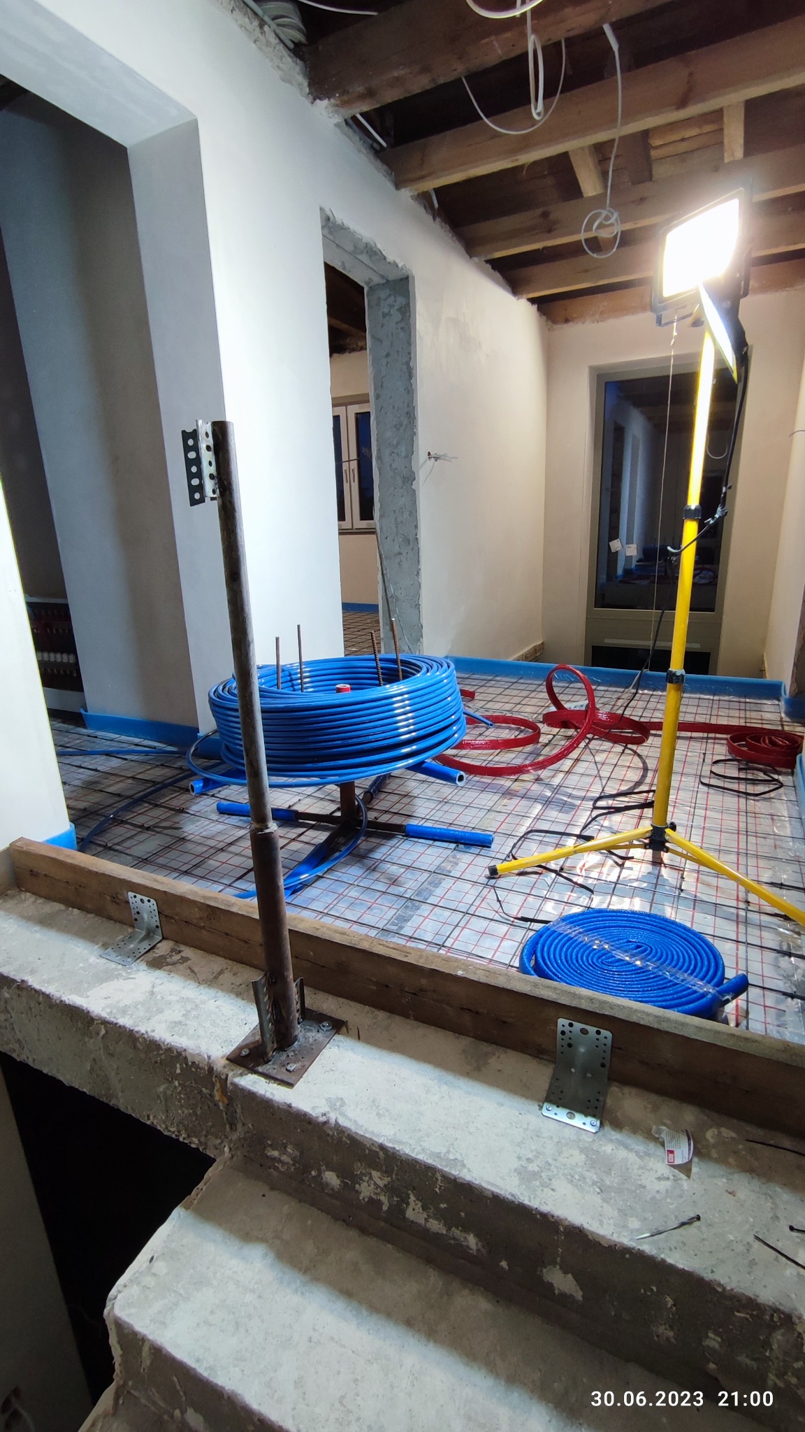 Niedokończona instalacja ogrzewania podłogowego z niebieskimi i czerwonymi rurami ułożonymi na siatce, oświetlona lampą budowlaną, widoczne elementy konstrukcyjne sufitu i podłogi.