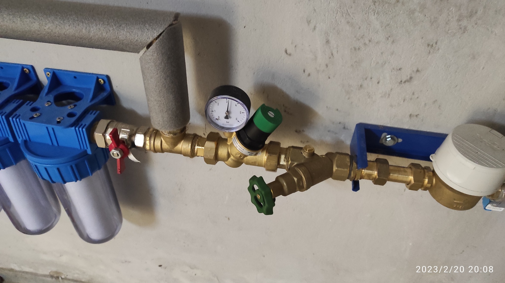 Fragment instalacji wodnej z filtrami, reduktorem ciśnienia z manometrem, zaworami kulowymi z zielonymi rączkami i wodomierzem, zamontowanej na szarej ścianie.