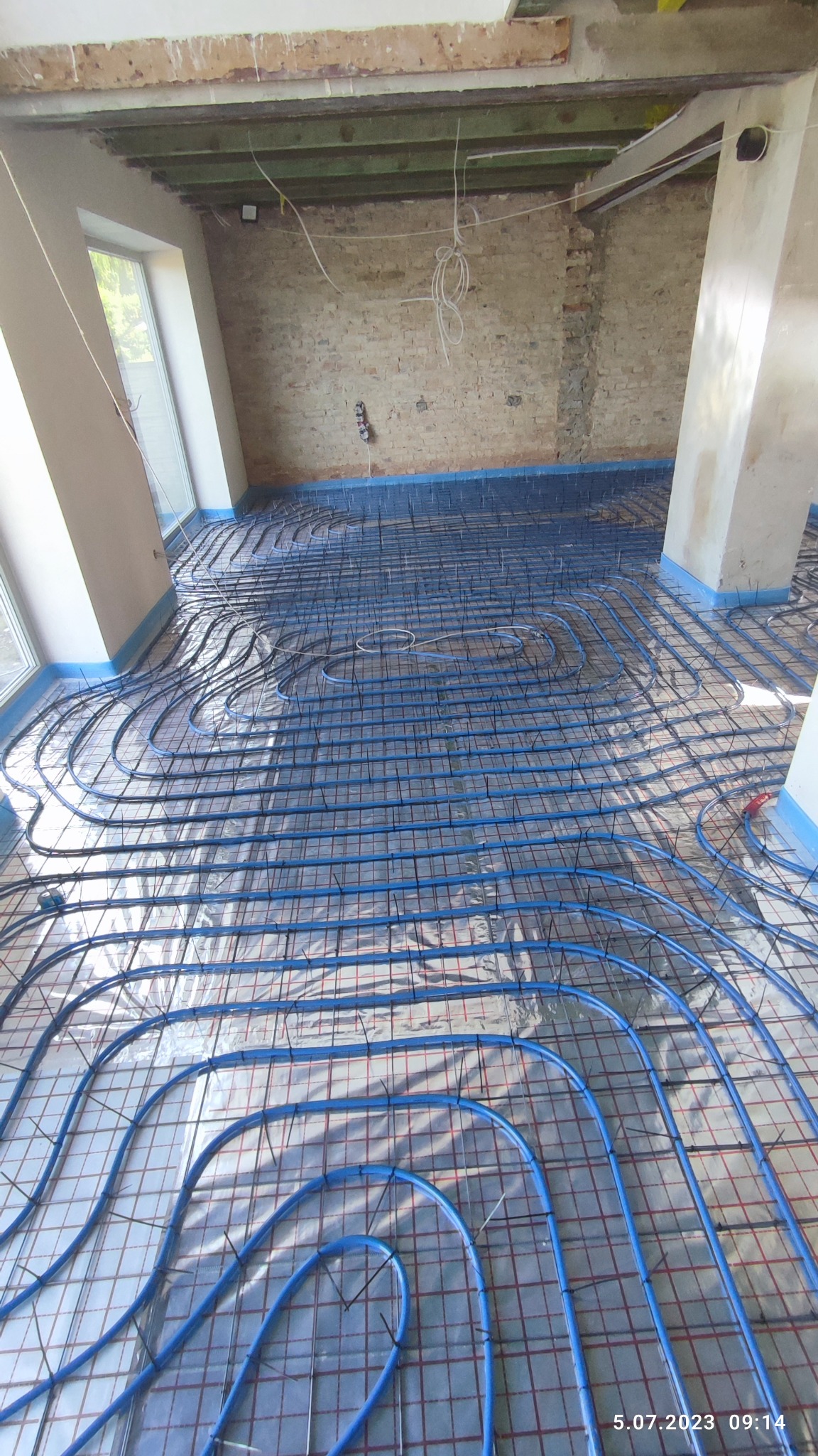 Niebieskie rurki ogrzewania podłogowego ułożone na srebrnej folii z czerwoną siatką na podłodze w surowym stanie, z widocznymi fragmentami ceglanej ściany i oknami.