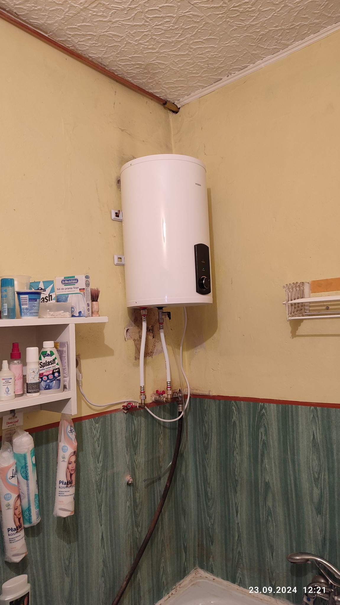 Biały, wiszący bojler elektryczny podłączony do instalacji wodnej w łazience o żółtych ścianach, widoczne rury doprowadzające wodę i półka z kosmetykami.