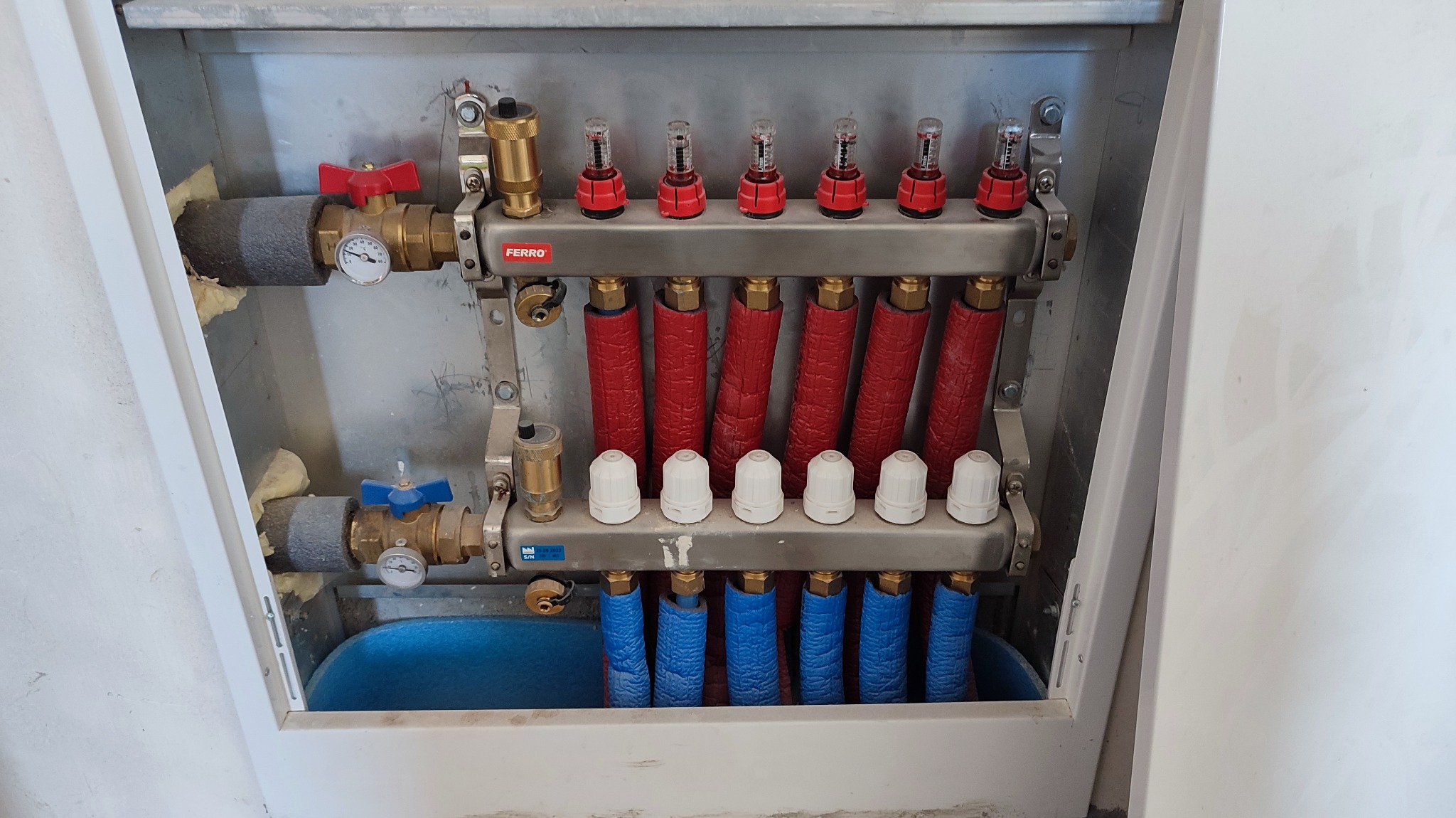 Rozdzielacz ogrzewania podłogowego w metalowej szafce, z czerwonymi i niebieskimi rurami, zaworami Ferro i manometrami, widoczne elementy instalacji w szarej izolacji.
