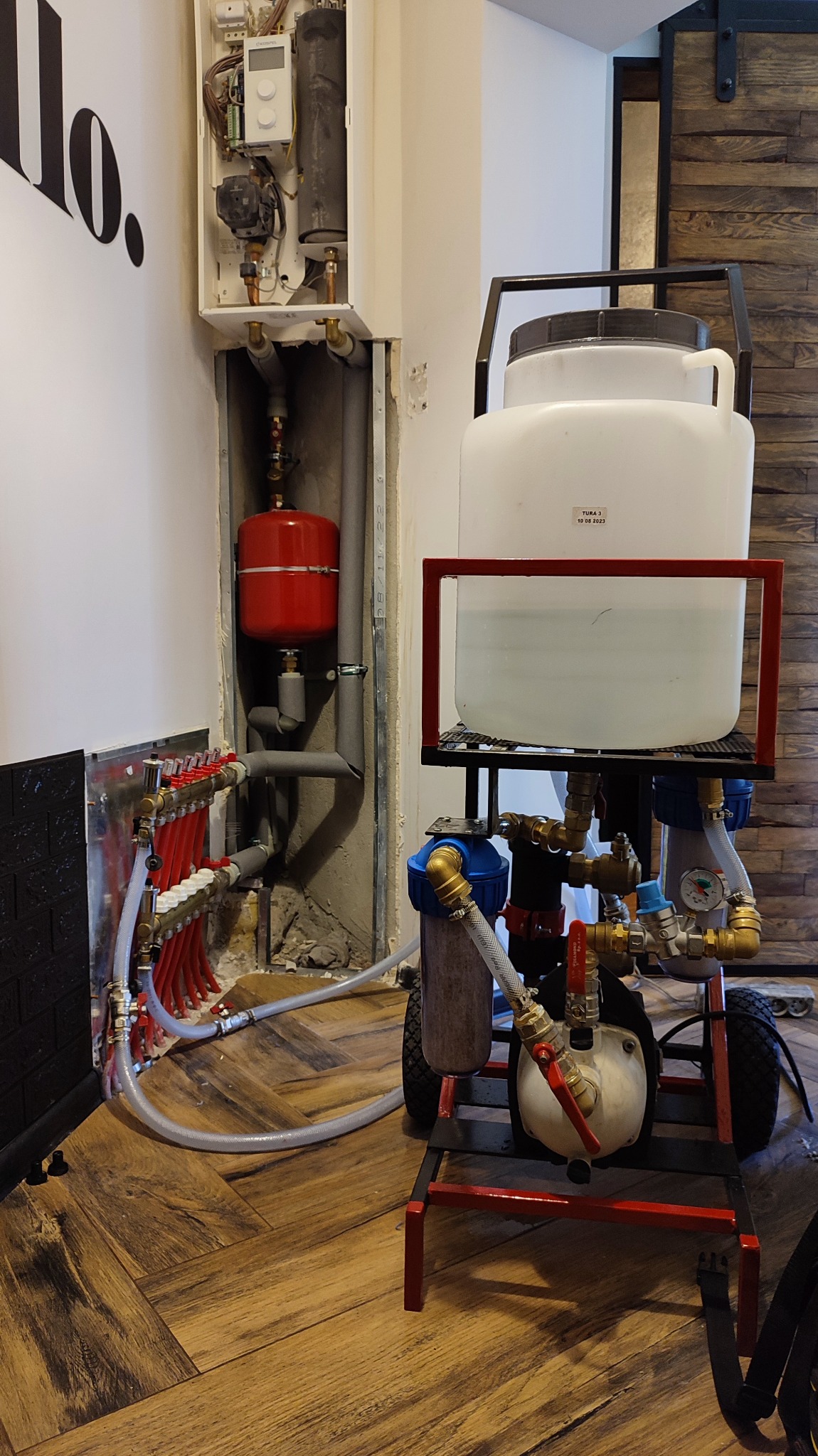 Instalacja grzewcza z rozdzielaczem, naczyniem przeponowym i kotłem w trakcie napełniania wodą za pomocą przenośnego zestawu do napełniania i odpowietrzania instalacji.