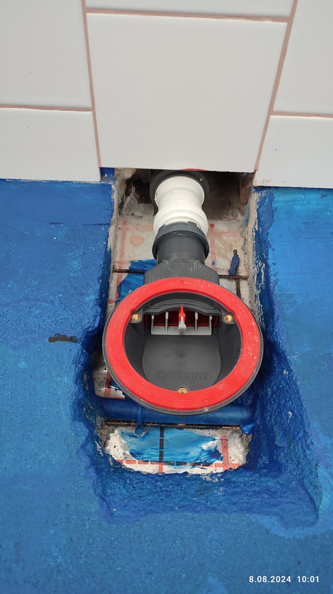 Montaż odpływu liniowego w łazience, widoczny syfon z czerwoną uszczelką, niebieska hydroizolacja na posadzce, białe płytki częściowo zamontowane na ścianie.