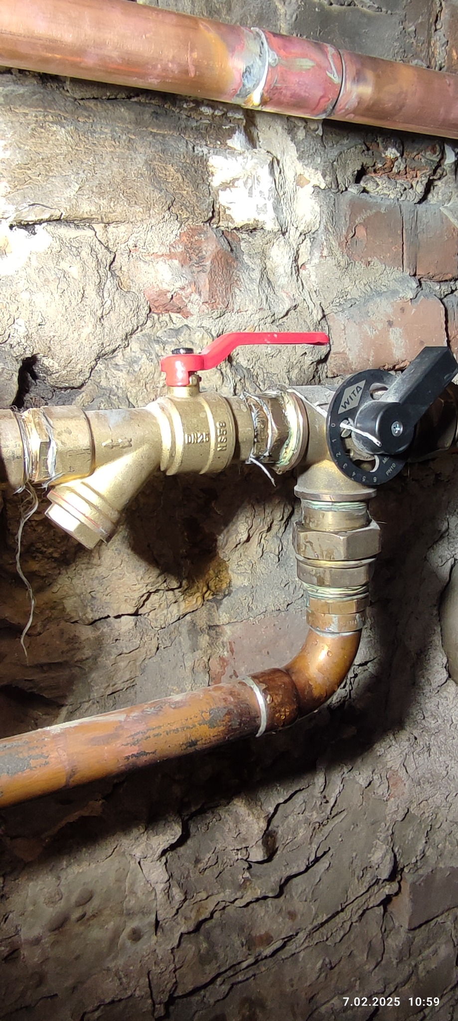 Mosiężny zawór z czerwoną rączką i czarnym pokrętłem Wita, połączony z miedzianymi rurami na tle surowej, kamiennej ściany piwnicy.