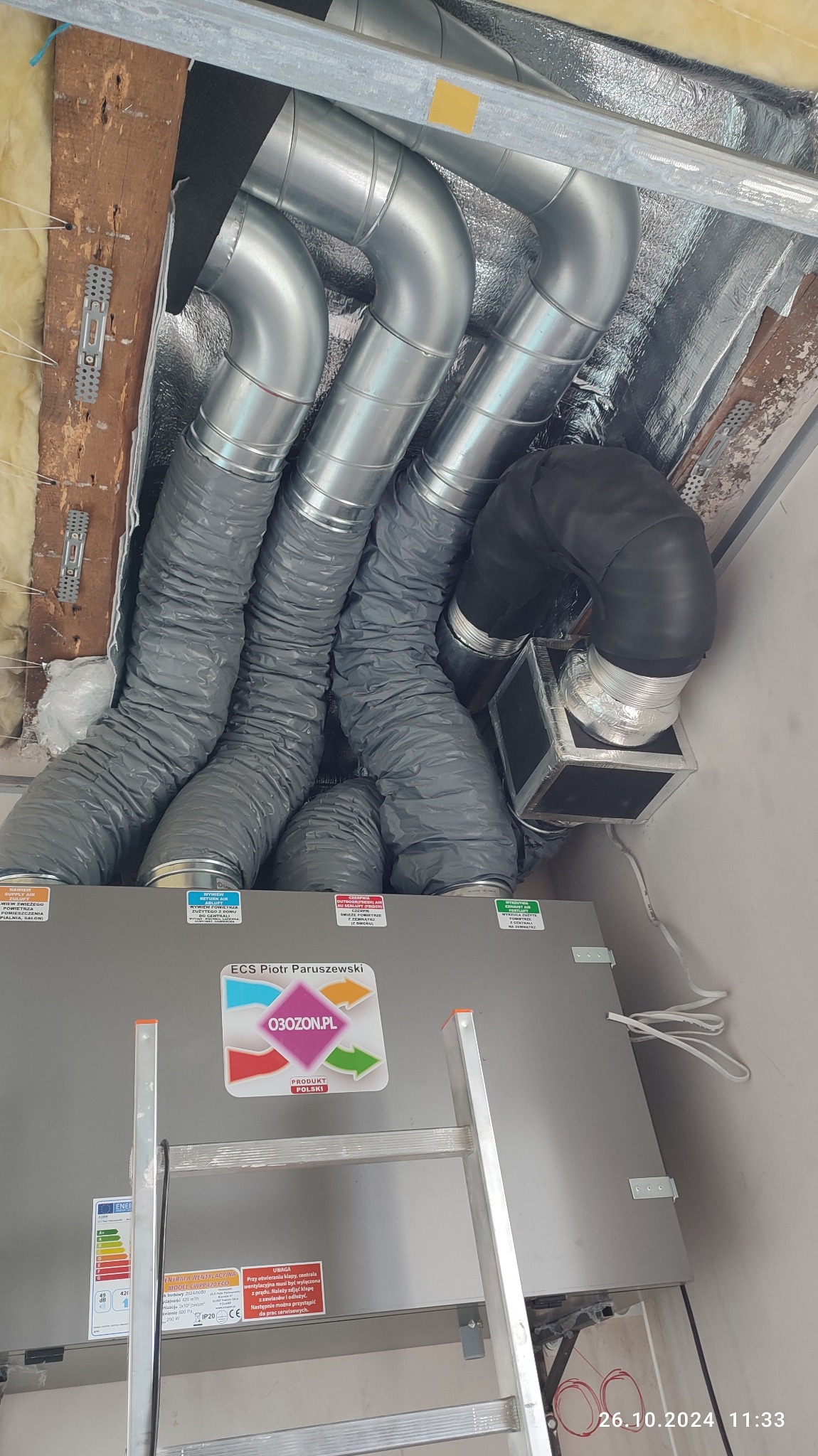 Centrala wentylacyjna z podłączonymi przewodami wentylacyjnymi, widoczne elastyczne rury szare i sztywne rury srebrne, instalacja w pomieszczeniu, drabina oparta o urządzenie.