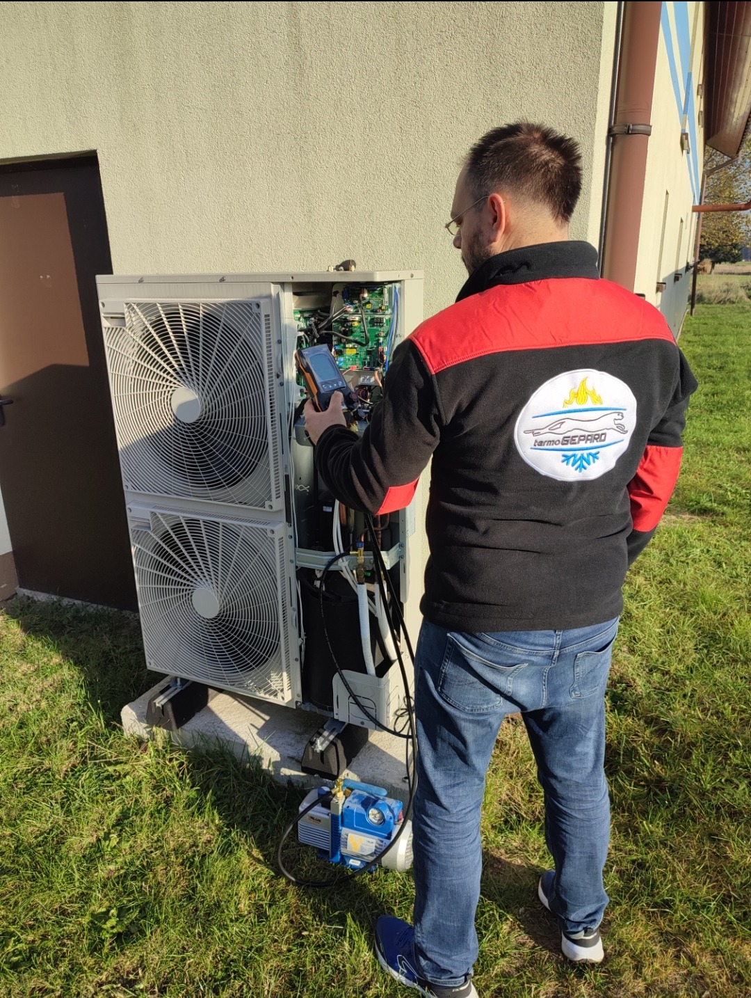 Instalator w okularach, w bluzie z logo firmy, mierzy parametry otwartej obudowy jednostki zewnętrznej klimatyzacji przy użyciu multimetru cyfrowego; obok stoi pompa próżniowa.