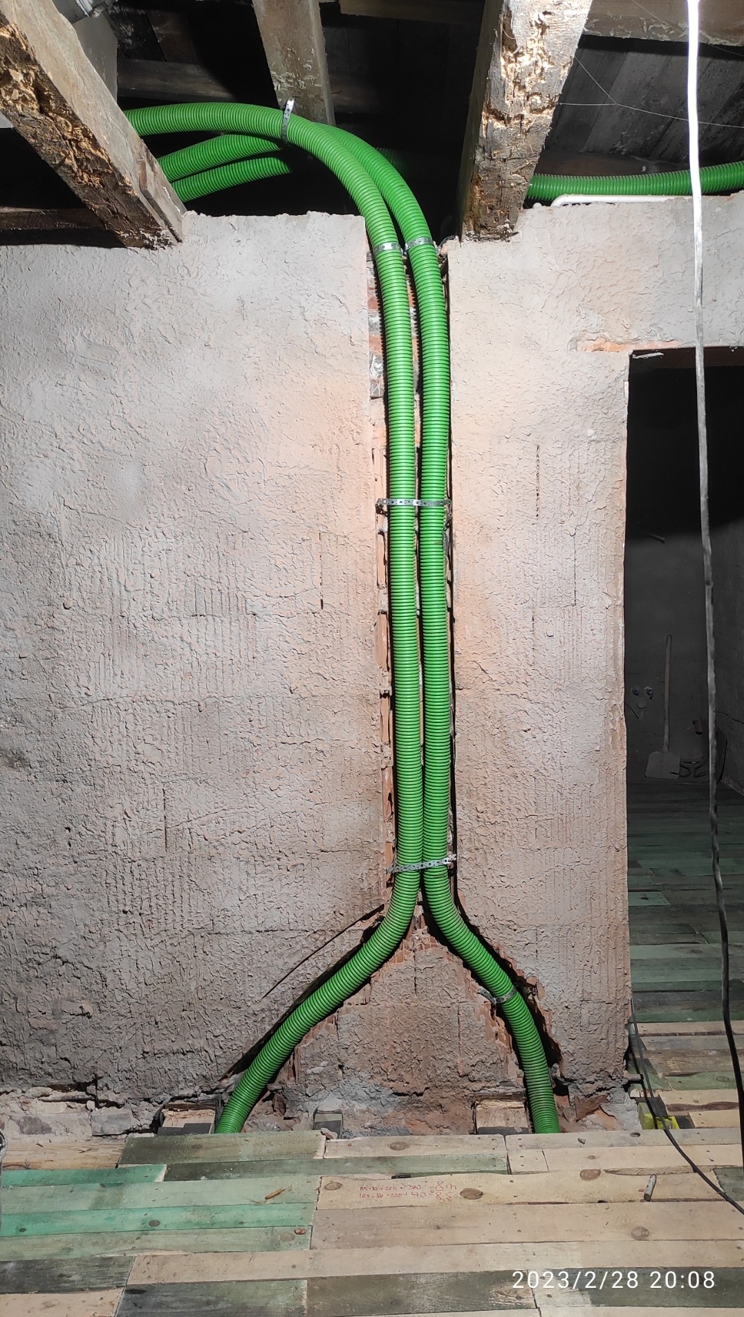 Dwie zielone, karbowane rury wentylacyjne przymocowane do surowej ściany z widocznymi śladami tynku i cegieł, przechodzące przez drewniany strop w trakcie instalacji.