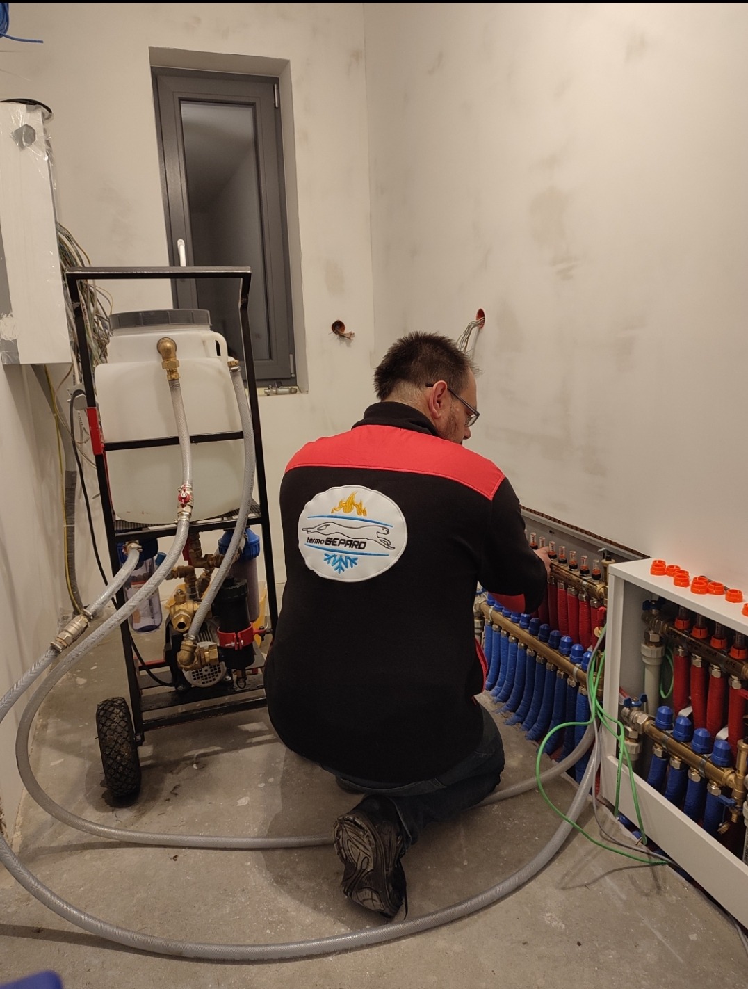 Instalator w Miasteczku Śląskim klęczy przed rozdzielaczem ogrzewania podłogowego, podłączając rury; widoczny wózek z pojemnikiem i pompą do napełniania instalacji.