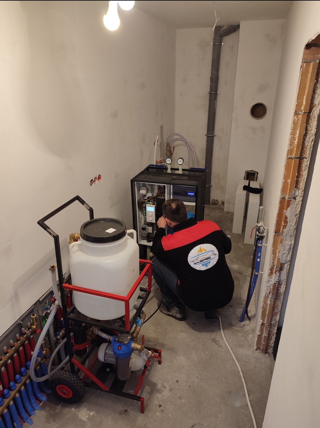 Instalator w czarno-czerwonej bluzie z logo firmy, w kucki, podłącza przewody do czarnej szafki sterowniczej w pomieszczeniu z widoczną instalacją grzewczą podłogową i rurą odpływową pod sufitem.