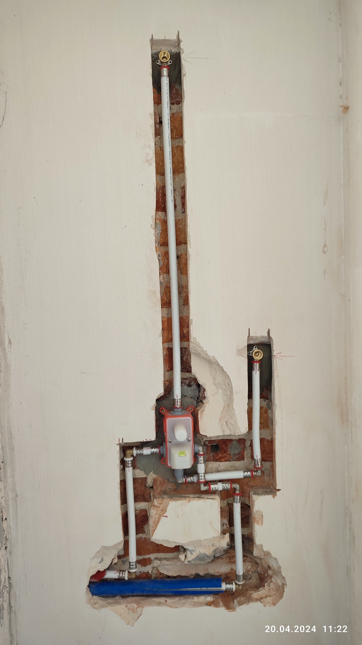 Fragment ściany z widoczną instalacją hydrauliczną w trakcie montażu: białe rury PEX z czerwonymi złączkami, niebieska rura, czerwona skrzynka z zaworem, odsłonięta cegła i fragmenty tynku.