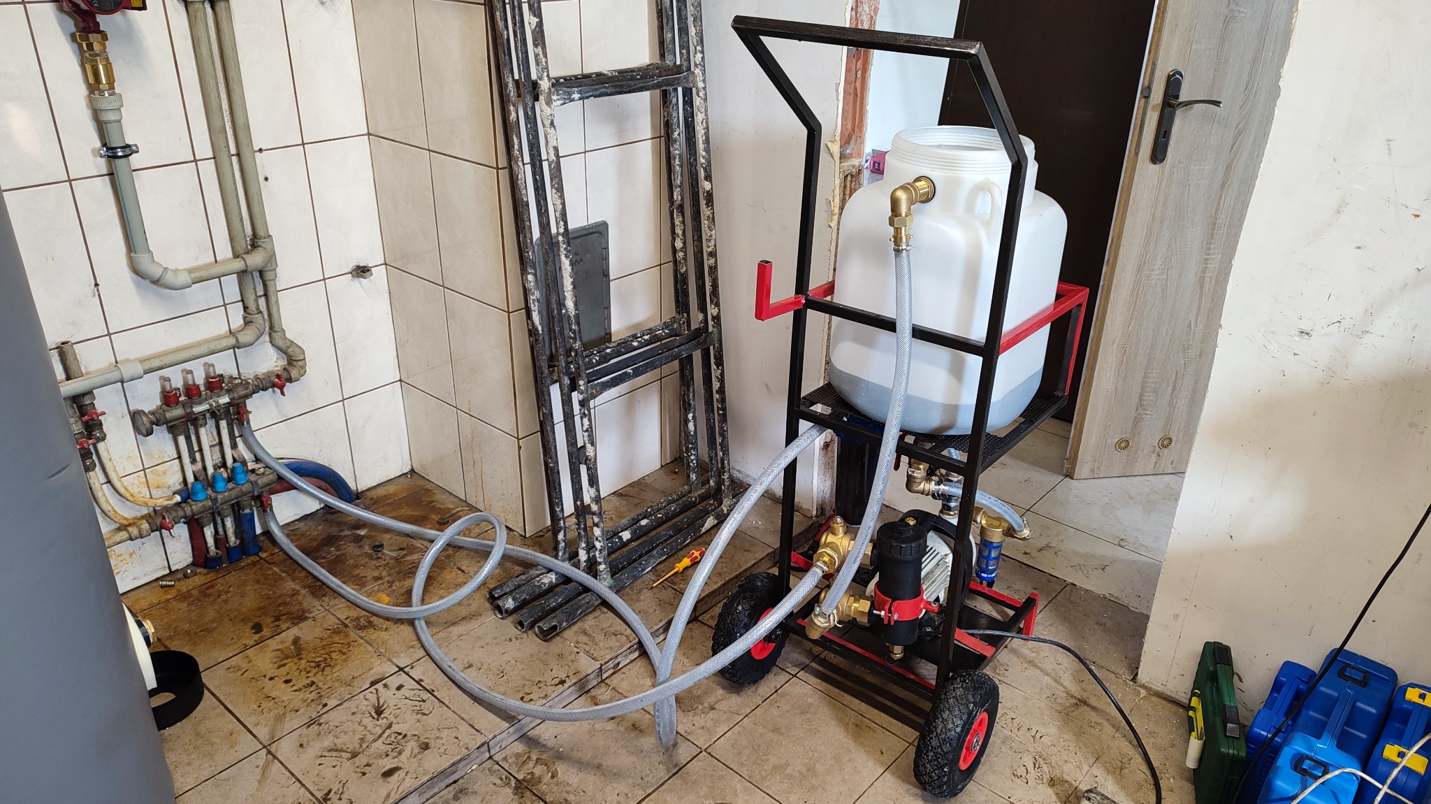 Przenośny zestaw do płukania instalacji grzewczej z plastikowym zbiornikiem na wózku, podłączony do rozdzielacza ogrzewania podłogowego w brudnej kotłowni z widocznymi rurami i drabinką.