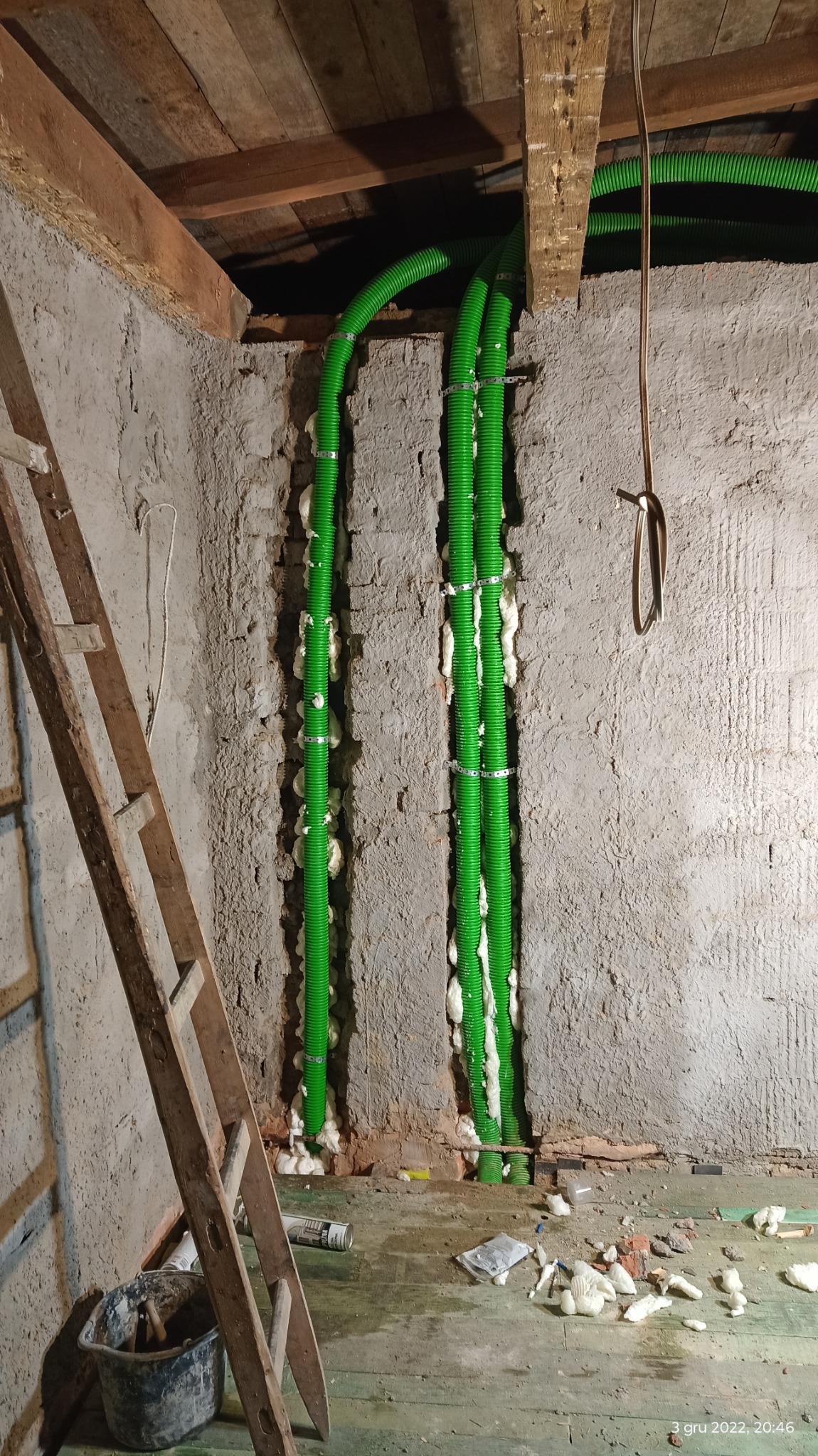 Montaż trzech zielonych rur karbowanych, przymocowanych metalowymi obejmami do ściany z surowego betonu, szczeliny wypełnione pianką montażową, widoczna drewniana drabina oparta o ścianę, podłoga...