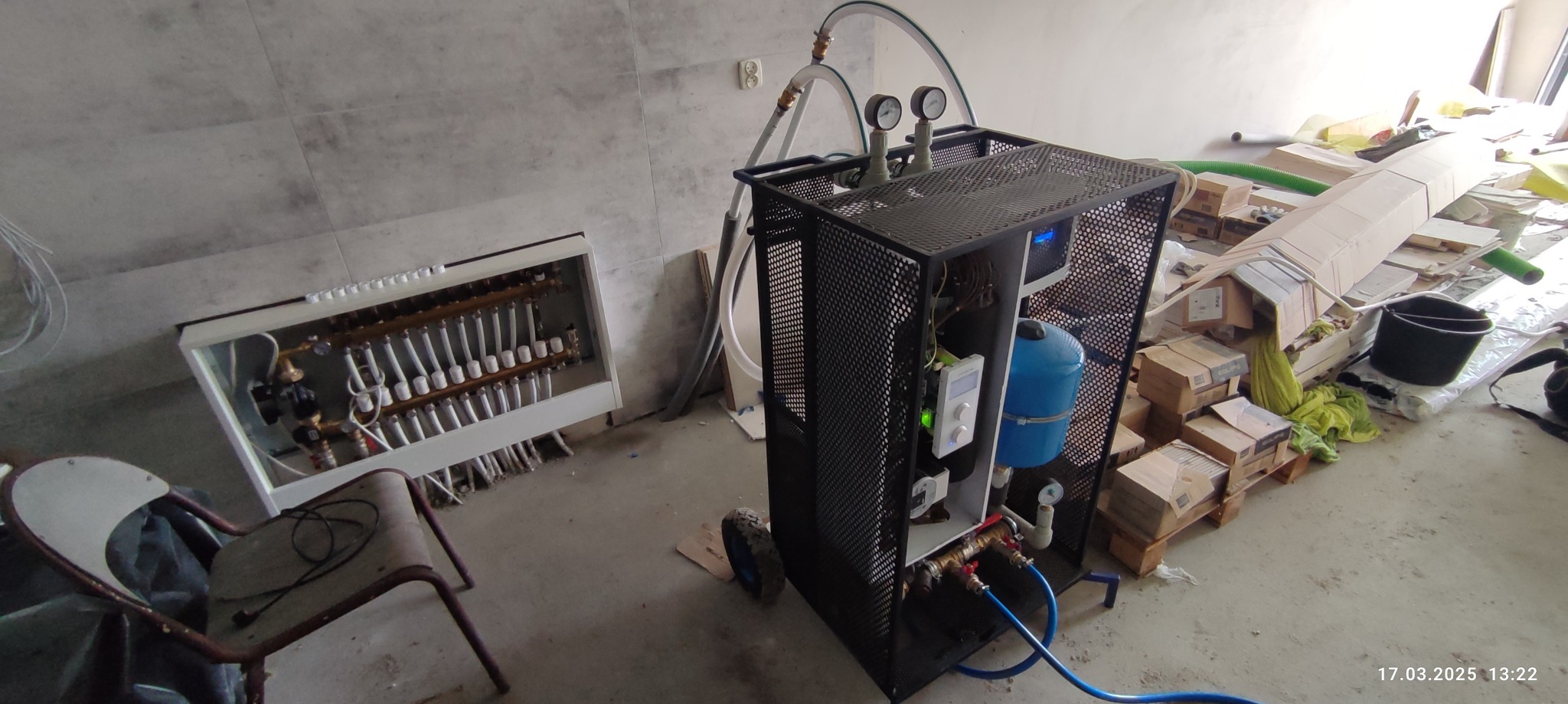 Rozdzielacz ogrzewania podłogowego w skrzynce, z podłączonym agregatem ciśnieniowym na kółkach, na tle surowego wnętrza z widocznymi materiałami budowlanymi.