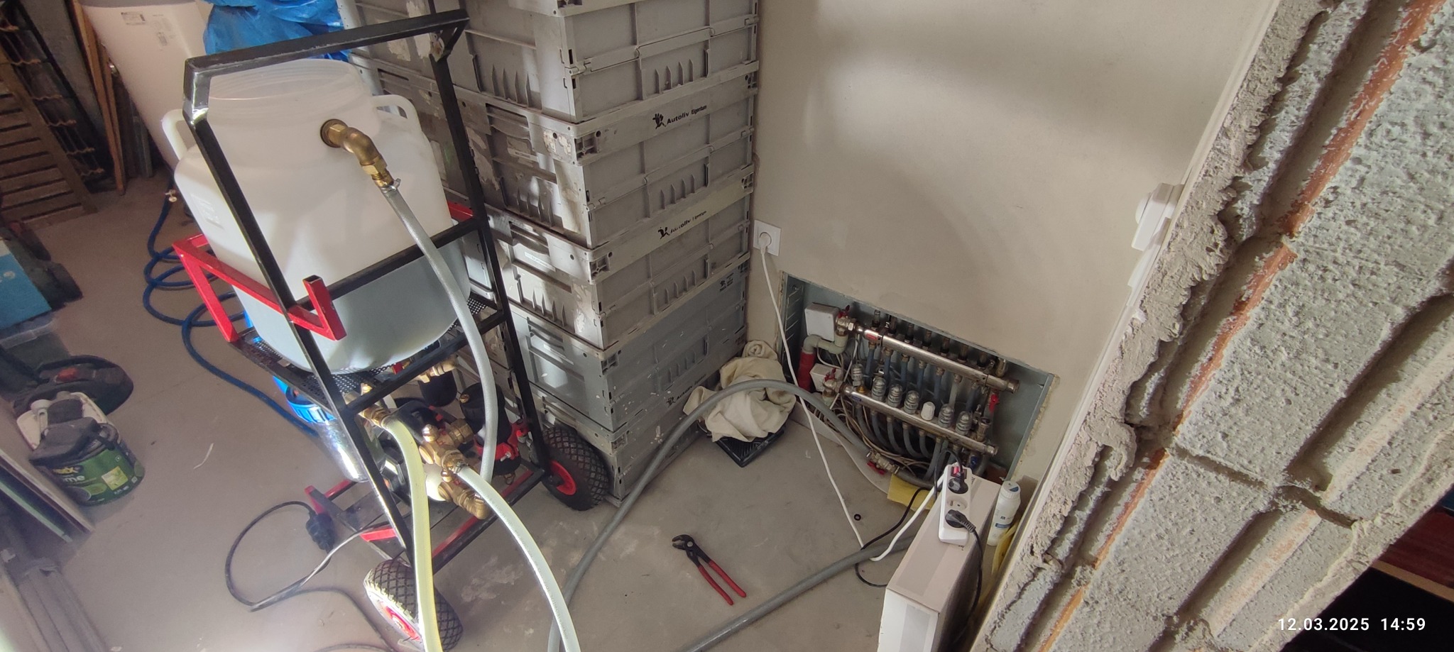 Próba ciśnieniowa instalacji grzewczej z użyciem mobilnego zestawu testowego, widoczny rozdzielacz wnękowy z podłączonymi rurami, sterta szarych skrzynek obok.