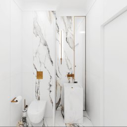 Anna Ostrowska - Minimalistyczna łazienka w bieli z akcentami złota: marmurowa ściana, wisząca toaleta, umywalka kolumnowa i złota armatura.