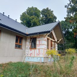 Techbud Piotr Podściański - Budowa domu szkieletowego: widok z zewnątrz na konstrukcję w trakcie ocieplania, z widoczną membraną i rusztowaniem. Dom z szarą blachodachówką, brązowe okna.